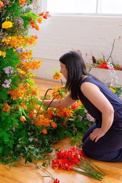 Florist Roxy kneeling on a wooden floor, carefully placing orange flowers into a vibrant, large-scale floral installation featuring marigolds, orchids, and greenery in a bright sunlit studio.