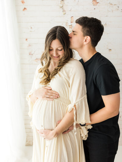 husband kissing pregnant wife in cream maternity gown for studio photoshoot