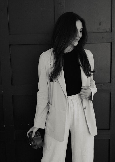 Reilly Erin smiling softly while holding her camera, dressed in a blazer and trousers, exuding warmth and professionalism.