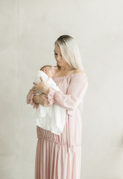 Mother in a blush pink dress holding her swaddled newborn against a minimal beige backdrop during a Kansas City newborn photography session.