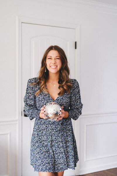 A warm, light-filled scene featuring Emylee Williams holding a disco ball in a stylish workspace — representing the balance of creativity and strategy she brings to her high-performing service provider clients.