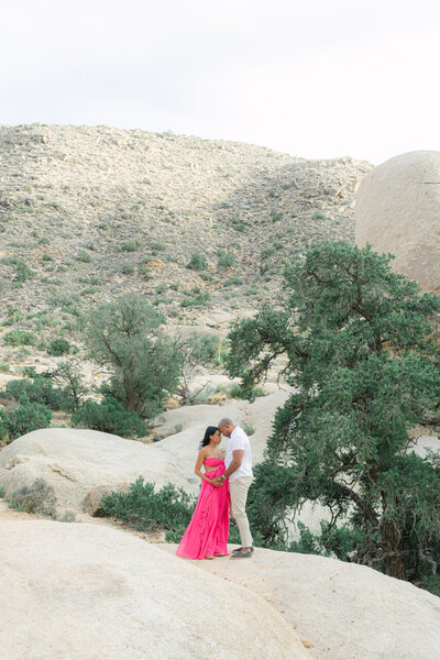 A mother-to-be in a vibrant pink gown, sharing a tender moment with her partner among the desert boulders of Joshua Tree National Park, captured in a refined editorial style by Elizabeth Kane Photography—serving Philadelphia, New York, and DC.