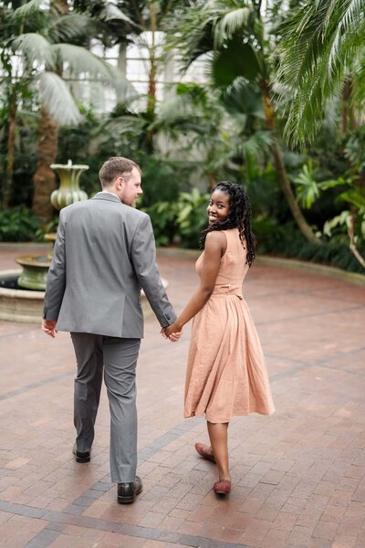 A young woman holds hands with her fiance and smiles back at the camera inside the Palm House