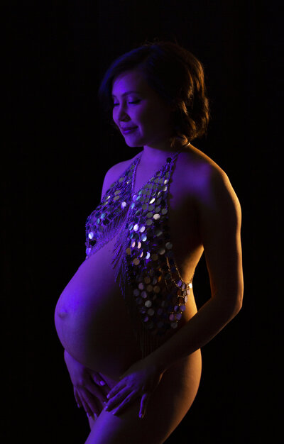 Charlotte creative Maternity Photograph created in our Fort Mill studio on a black backdrop with purple and yellow gel lighting with mom wearing a silver sparkle top while mother to be of asian decent smiles at her belly