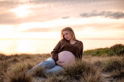 Close up of a pregnant belly with the mother wearing a brown jumper and jeans while leaning against a white weatherboard wall