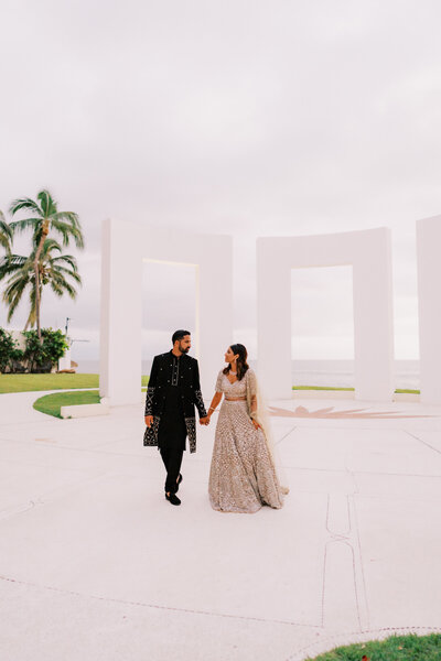 A couple dressed in traditional Indian wedding attire walk hand in hand in an open-air architectural setting with large white columns. The groom is wearing a black sherwani, and the bride is in an ornate light-colored lehenga. Keywords: destination wedding planning, luxury destination weddings, traditional Indian wedding.