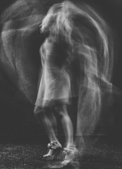 Abstract black and white photo of a woman in a short dress and heels, captured in motion blur with flowing, ghostlike trails surrounding her.