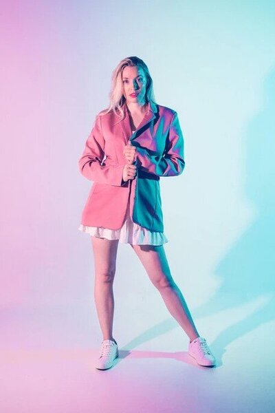 A woman with blonde hair and bright lipstick wears a pink blazer over a white dress in front of a pastel background