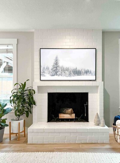 white brick fireplace with tv above, beige walls and large window, two potted plants with large green leaves
