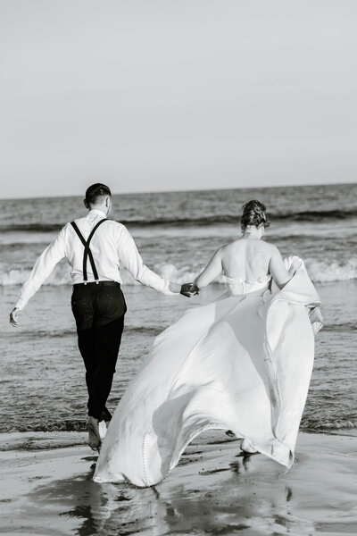 Rhode Island Elopement | A couple in wedding attire walks hand in hand along the beach, their backs to the camera. The bride's dress billows in the breeze.