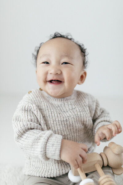 toddler smiles at the camera whilst holding a little wooden toy in the studio during his photoshoot