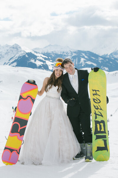 Bride and groom in wedding attire posing with colorful snowboards on the snowy slopes of Kitzbühel, Austria — playful after-wedding ski session by Studio Rose