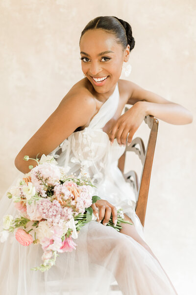 Bride poses for a formal magazine-worthy bridal portrait. She is sitting against a textured backdrop on a vintage chair. She is sitting sideways on the chair with one arm draped casually across the back. her florals are resting gently on her lap. The bride is looking over her shoulder and smiling at the camera.