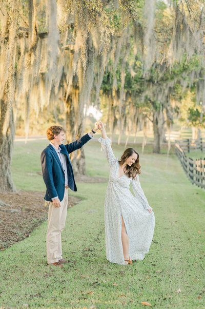 Engaged couple standing under a glowy tree while man holds womans hand as she spions
