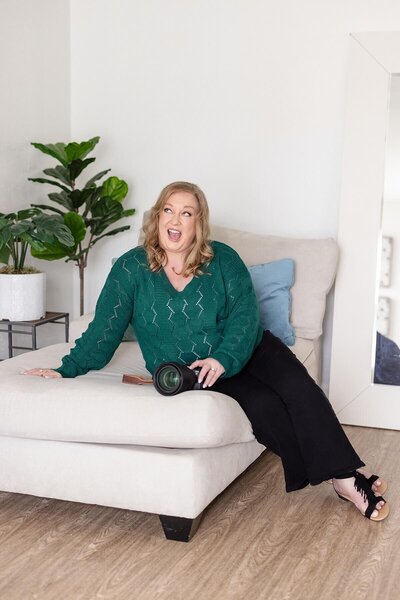 Megan Sethia, Seattle brand photographer and founder of She Plays with Fire, sitting on a modern cream chaise lounge in a lifestyle studio, holding her camera and laughing with personality and confidence.