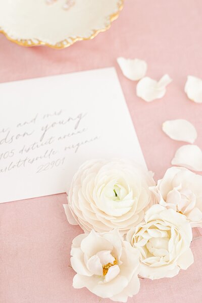 wedding invitation styled with a string of petals and a few ranunculus blooms on a pastel pink background.