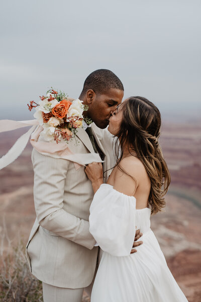 Utah elopement photographer captures man and woman kissing on top of a red rock mountains in Utah for wedding portraits