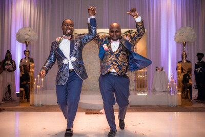 Gay african american couple jumps over broom after wedding ceremony. Dressed in floral accented gold and blue suits with pink accents. Ceremony backdrop features and gold boarder and white rose flower wall.