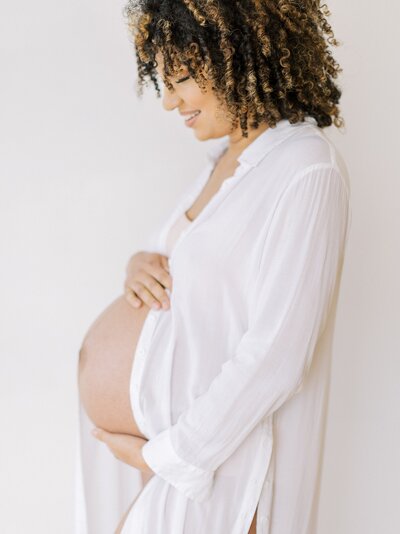 Side view of a pregnant woman wearing a long white shirt looking down and her baby bump