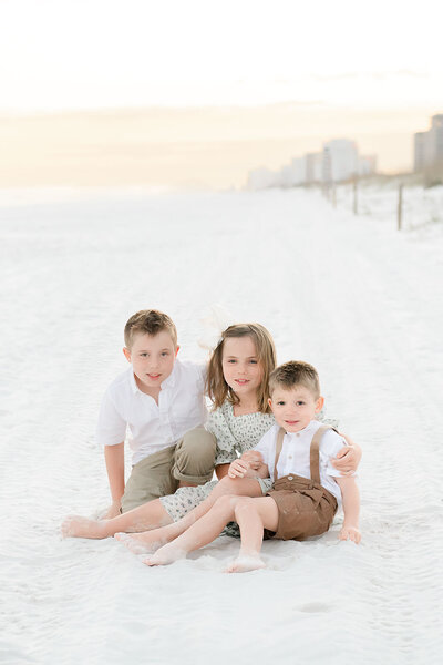 Three children cuddle together on the sand at sunrise, capturing a playful sibling moment with pastel skies and beach views.