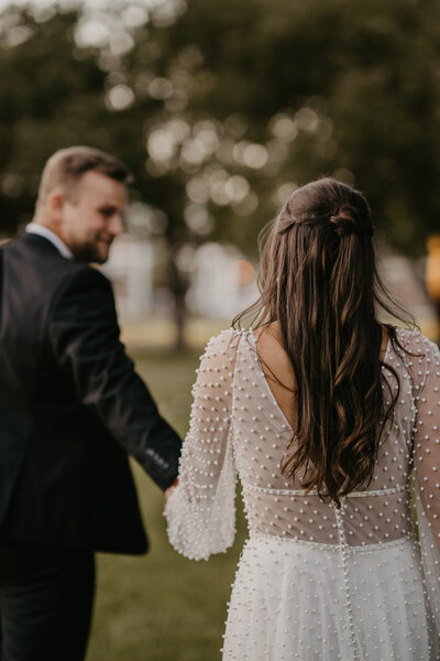 Bride and groom walk away from camera as groom is blurred out, but smiling back at his beloved wife. Wife's face cannot be seen but the camera focuses on her beautiful brown hair and gorgeous dress with pearls.