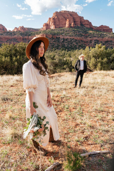 Bride in vintage western dress and wide-brim hat holding a bouquet during her Sedona elopement at Bear Mountain, with the groom standing in the distance.