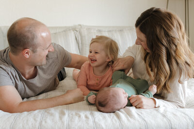 Longmont family sitting with newborn on bed while toddler is laughing and smiling at dad