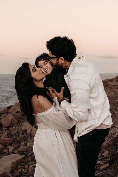 A couple stands outdoors by rocky terrain, each kissing a laughing child they hold together, against a backdrop of a serene sunset sky captured beautifully by an NSW family photographer.