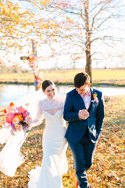 Bride and groom smiling and walking hand in hand outside with yellow leaves on the ground