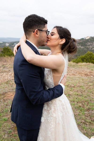 An Austin-based wedding photographer captures a romantic moment between a bride and groom as they share a passionate kiss in a scenic field with majestic mountains serving as the breathtaking backdrop.