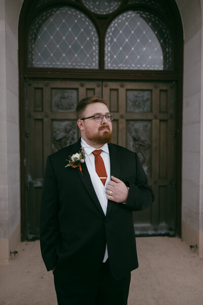 Wedding Photography of groom, looking out into distance with his hand on his suit jacket, wearing an orange tie and glasses standing in front of a vintage looking church door in Milwaukee, WI, captured by Morgan Ashley Lynn Photography