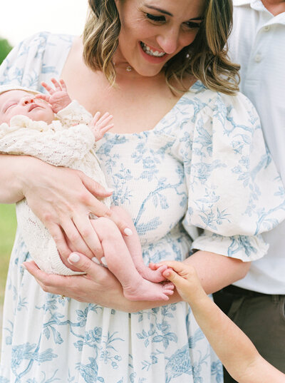 Brunette mother holds newborn daughter as toddler daughter's hand sneaks into frame to hold the baby's foot