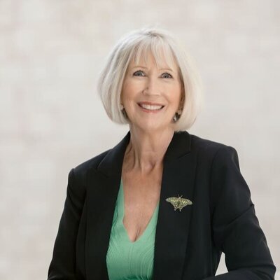 A woman with short, silver-blonde hair smiles warmly at the camera. She is wearing a black blazer with a butterfly brooch and a green top, with a light, neutral background.