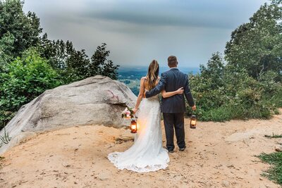 A bride and groom stand looking out at a mountain overlook after eloping. Their arms are wrapped around each other and their backs face the camera. They are standing on a dirt clearing with a large rock nearby and lush green trees. The bride is wearing a fitted white wedding gown and the groom is wearing a navy blue suit.