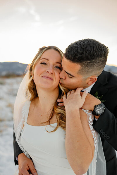 Romantic sunset photo of a newlywed couple with groom kissing his brides cheek. Scenic Colorado Springs mountains in the background, taken by Michelle Betz Photography, a top wedding photographer in Colorado Springs