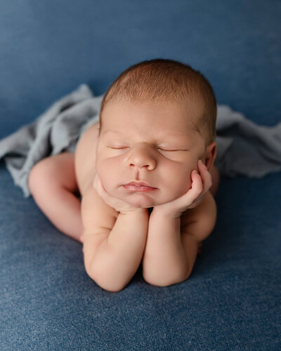 Peaceful baby boy resting with his chin in his hands in the froggy pose, styled on a blue backdrop with soft wrap details. Photographed by Inland Empire newborn photographer Kaitlyn Dawn.