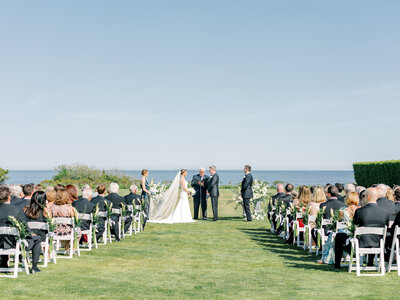 Bride and groom at the altar of their outdoor wedding ceremony in front of the water with guests in white chairs