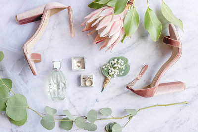 Flatlay image of wedding day accessories like bridal shoes, perfume, rings, boutonniere, and protea flower,