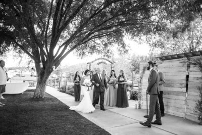 Father walking bride down aisle at wedding under a big tree. Bridesmaids wait in the background holding their flower bouquets