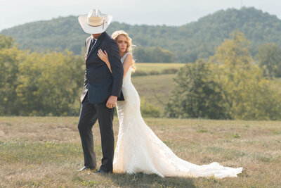 A Kentucky bride and groom pose for a portrait after their backyard wedding at the groom's family farm. The groom is wearing a cowboy hat and looking down while the bride hugs him from behind and looks the other direction.
