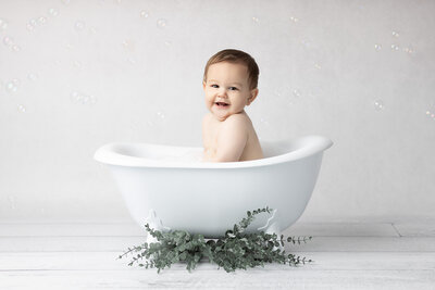 Smiling baby boy in a white clawfoot tub surrounded by bubbles and greenery on a whitewashed wood floor, captured by a Dallas milestone photographer.