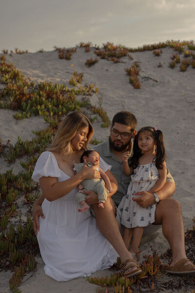 Family sitting together on the beach with their newborn and toddler, enjoying a sunset moment