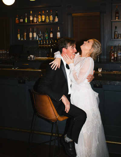 Flash photo of groom sitting in a chair while bride has her arms around him in front of a bar