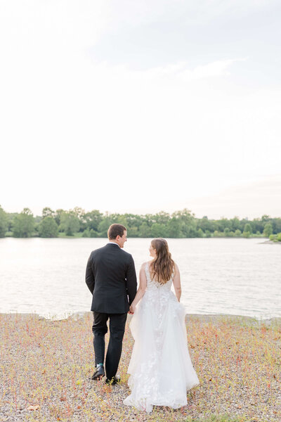 A bride and groom walk on the beach toward the lake looking at each other.