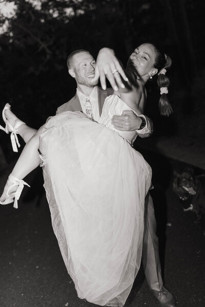 Groom carrying his bride down the road while she laughs and shows off her ring, captured by a Spokane wedding photographer.
