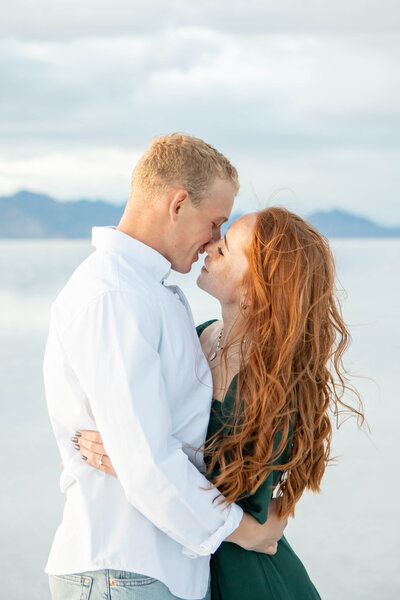 An engaged couple hugs and looks at each other at the Bonneville Salt Flats Utah. He's wearing a white shirt and she's in a green dress. Her red hair blows in the wind.