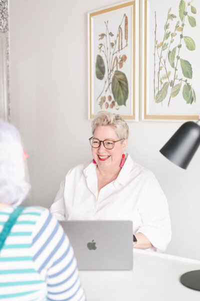 In a bright office, Pippa, a smiling person with short blond hair, glasses, and hot-pink tassel earrings sits in a white shirt behind a laptop, chatting with someone in the foreground; a black desk lamp and framed botanical prints hang on the wall.