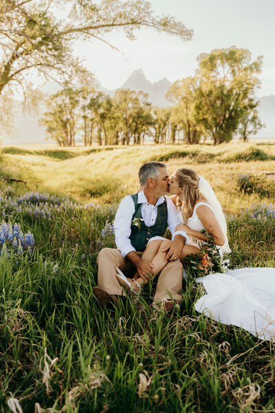 bride and groom sitting on the ground kissing in a field with mountains in the background