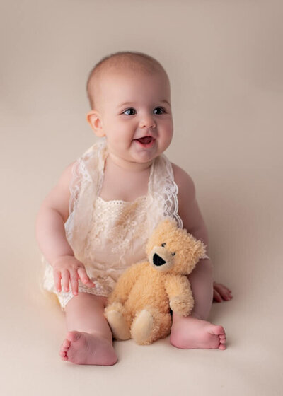 6 month old girl in a lace romper on a beige backdrop and a teddy bear in her lap