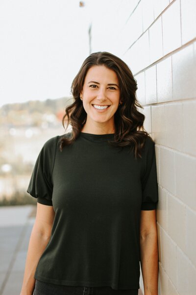 A woman named Roxanne Sardone in a black top leans against a wall, looking relaxed and contemplative.